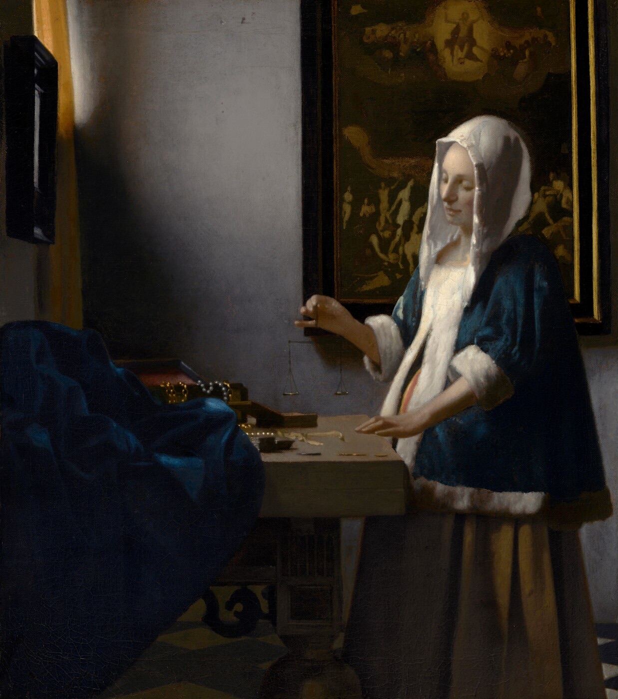 A woman in a blue jacket and white head covering holds a small balance beside a table in a quiet room.